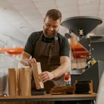 Business owner weighs paper bag with coffee beans on a scale at coffee factory